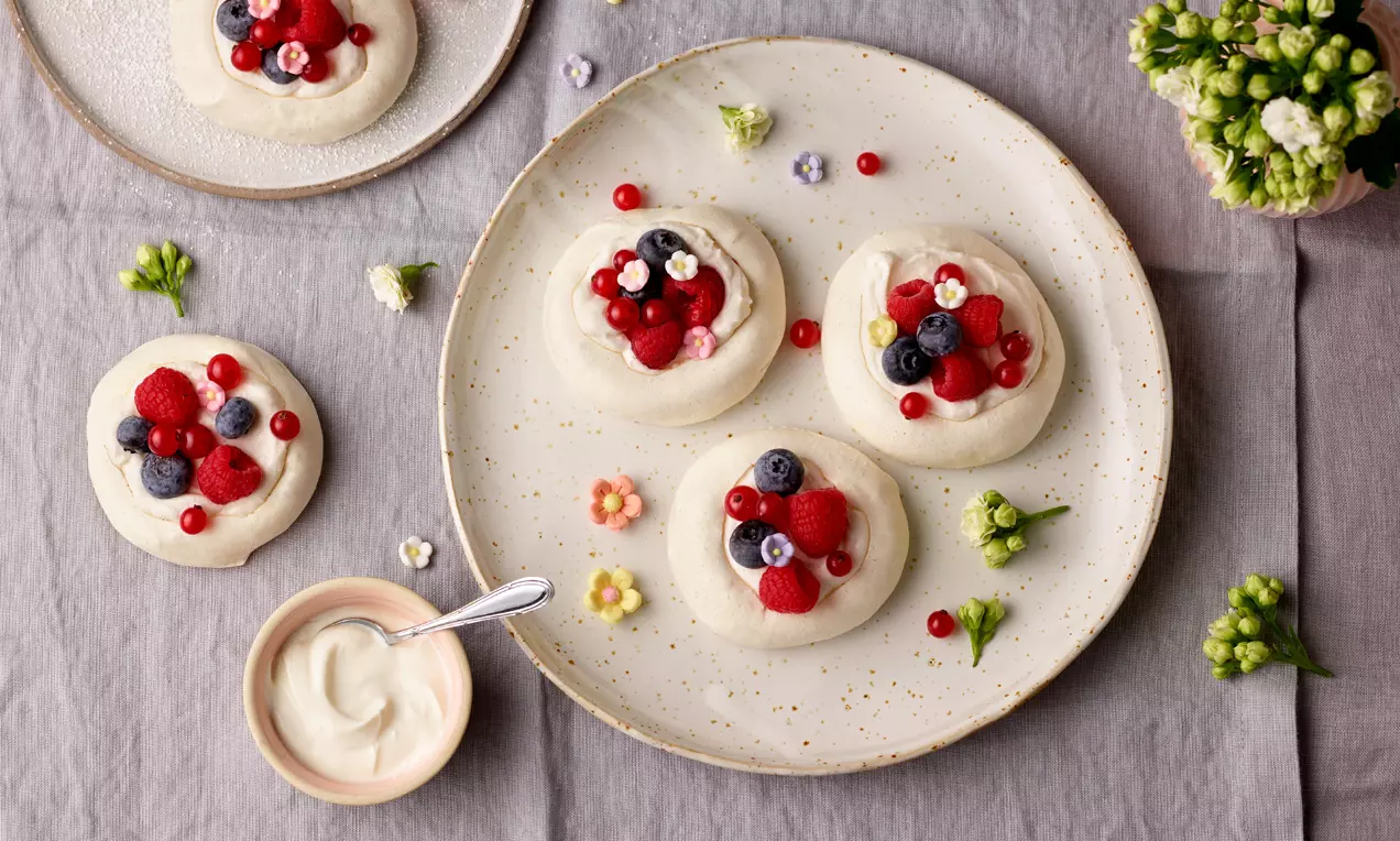 Mini Pavlova s bijelom, glatkom podlogom od bezea, ukrašena sočnim crvenim i plavim bobičastim voćem na neutralnom tanjuru.