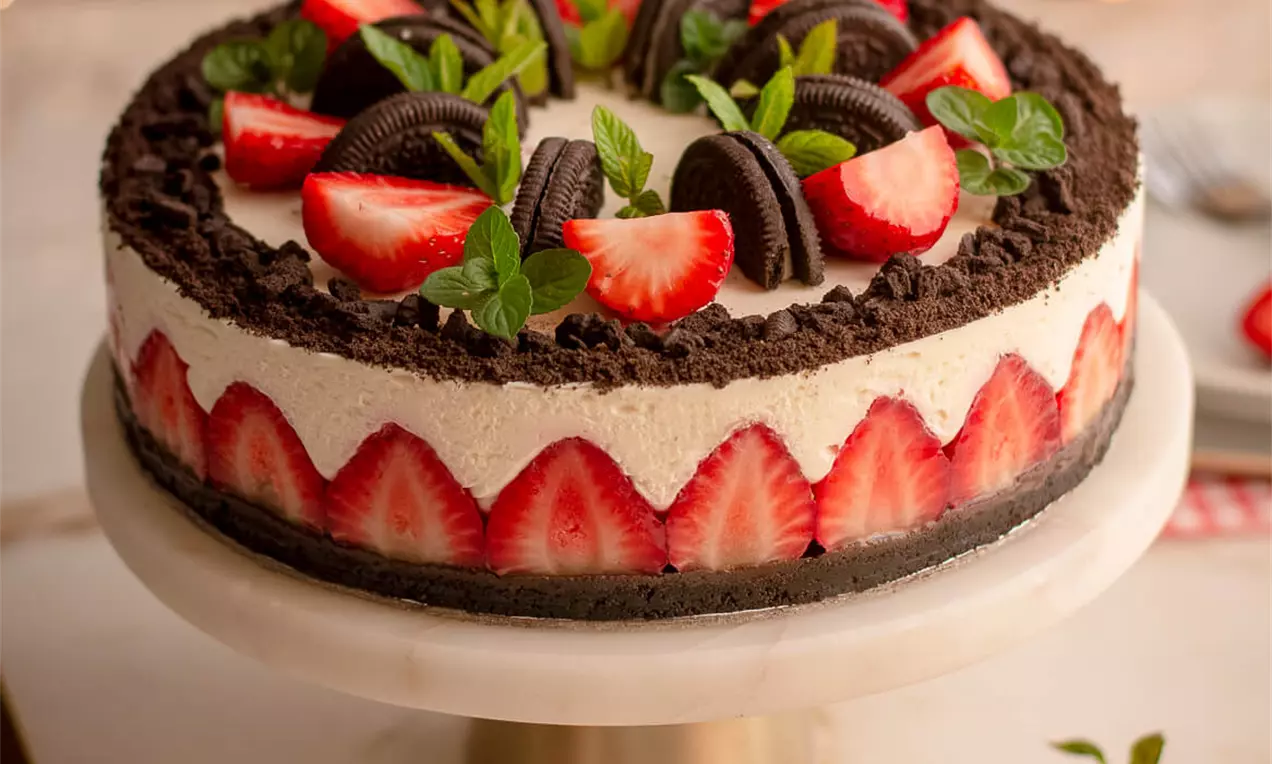 Torta na tamnoj podlozi od keksa s kremastim bijelim slojem, ukrašena jagodama, Oreo keksima, mentom i rubno postavljenim jagodama