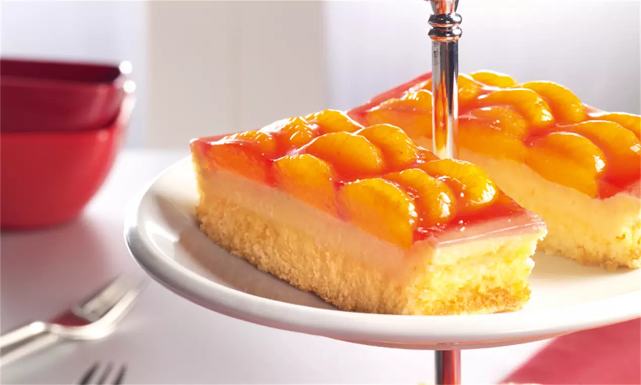 Dvije pravougaone kriške svijetložutog biskvita prekrivene laganom kremom i sjajnim slojem mandarina na bijelom tanjiru