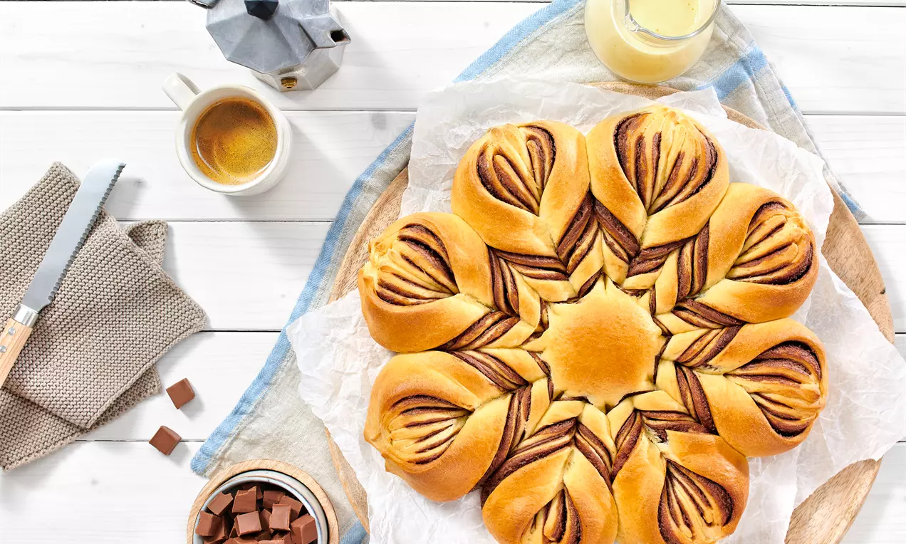 Zlatnosmeđi cvjetast oblik s uvrnutim listovima punjenim tamnom čokoladnom kremom na drvenoj podlozi s bijelim papirom