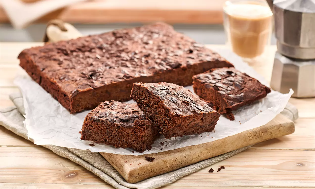Kvadratni čokoladni brownies s grubljom teksturom, posluženi na papiru za pečenje na drvenoj dasci uz šalicu kave i metalnu džezvu