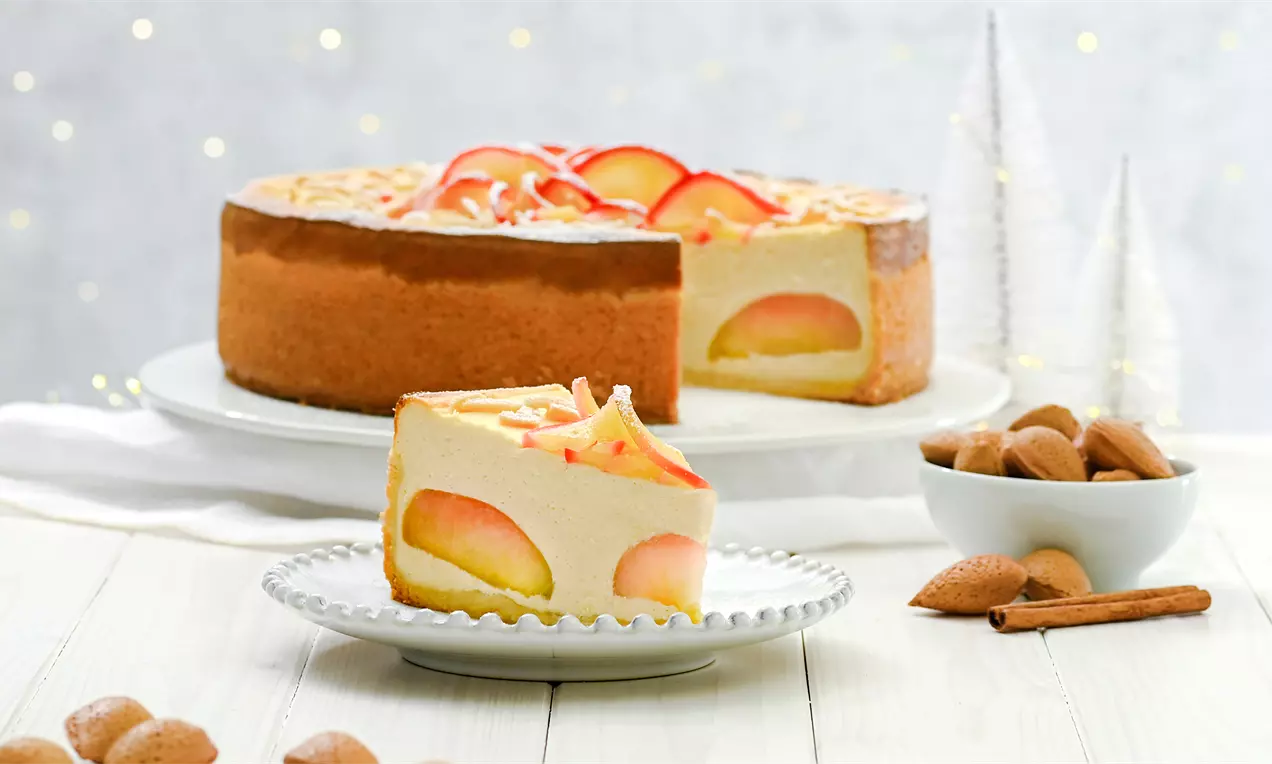 Slojevita torta svijetložute kremaste teksture s polovicama pečenih jabuka, ukrašena tankim crvenim kriškama na bijelom tanjiru