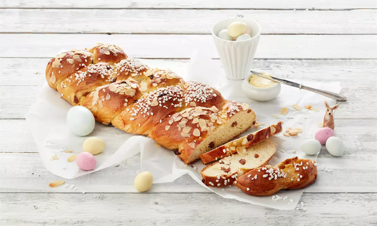 Pletenica od svijetlosmeđeg tijesta sjajne površine, posuta bijelim krupnim šećerom i listićima badema, na bijelom papiru uz šarena jaja