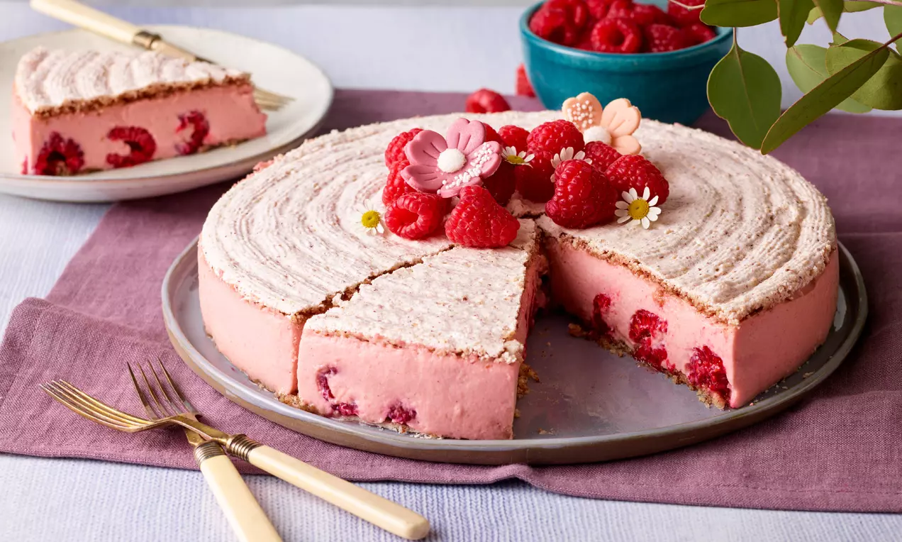 Svijetloružičasta torta s kremom od maline, ukrašena svježim malinama, cvjetićima i prhkom koricom na tanjuru s ljubičastim podloškom