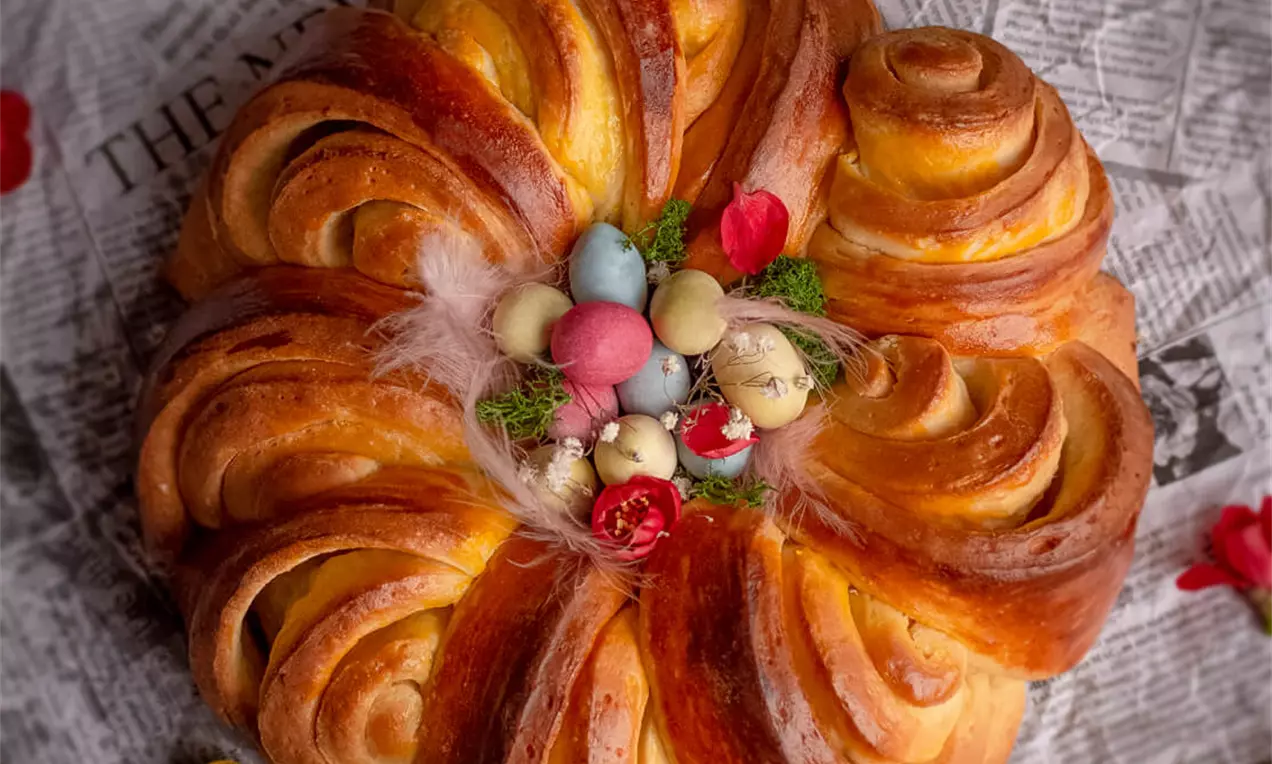 Blago zapečeno, zlatnosmeđe pleteno pecivo kružnog oblika s glazurom, ukrašeno obojenim jajima i cvijećem na podlozi od papira