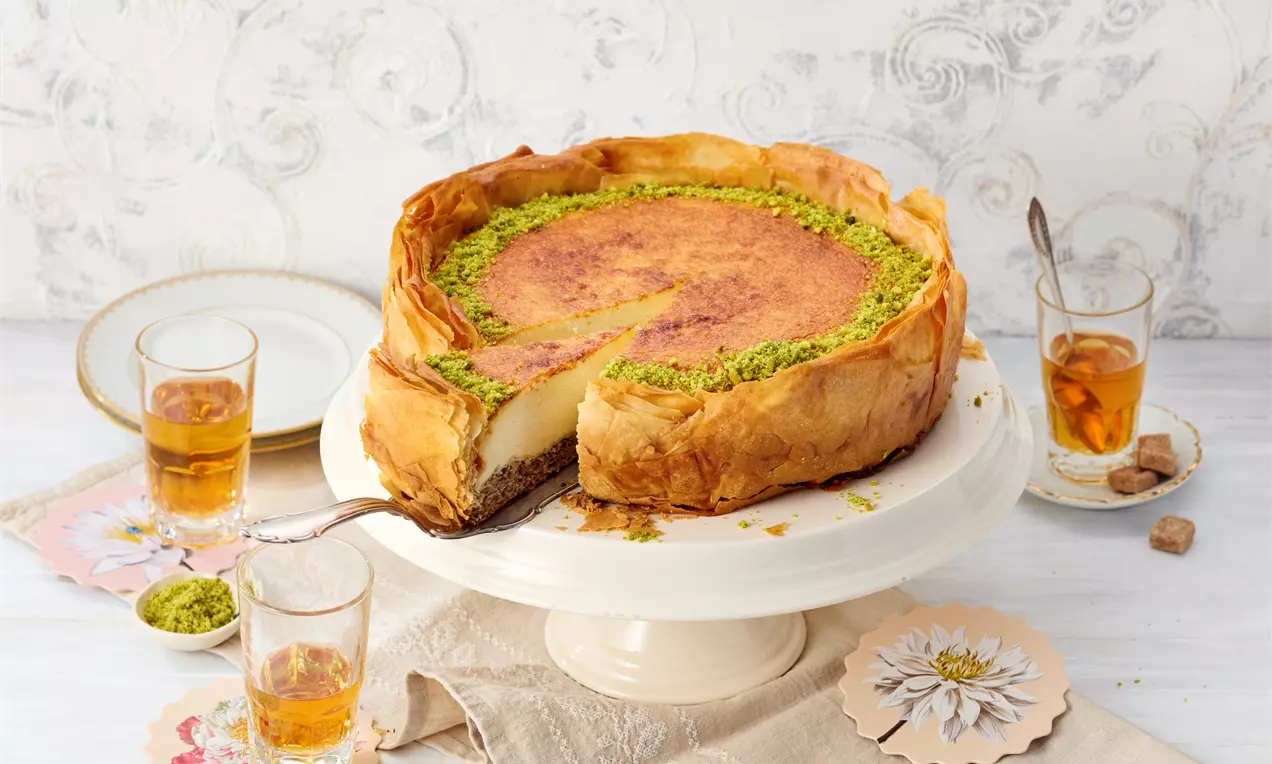 Baklava cheesecake na bijelom postolju, sa slojevima zlatnohrskave kore, kremastim punjenjem i preljevom od pistacija uz čaše čaja