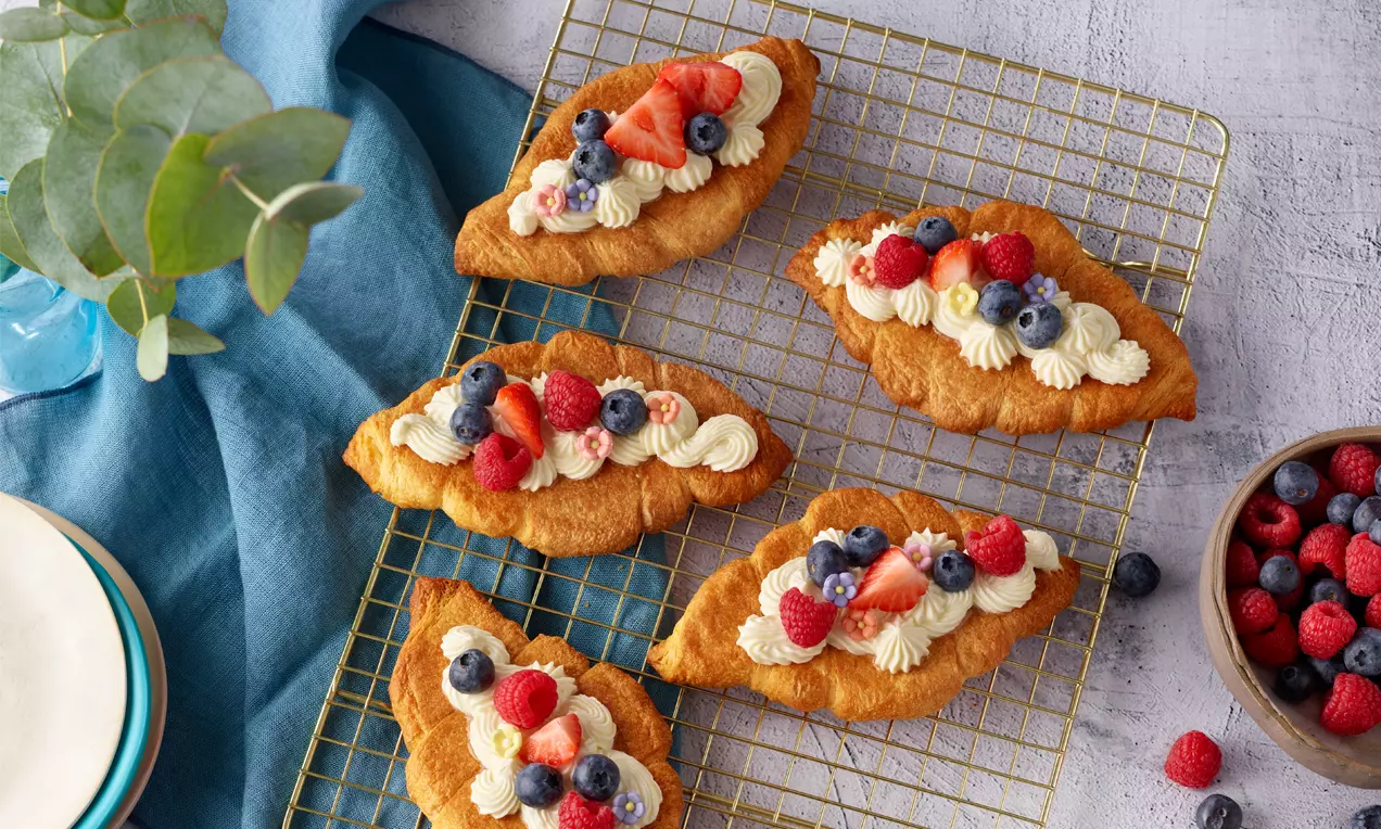 Zlatnosmeđi kroasani u obliku čamca na rešetki, ukrašeni svježim šlagom, jagodama, malinama i borovnicama, uz zdjelu bobičastog voća