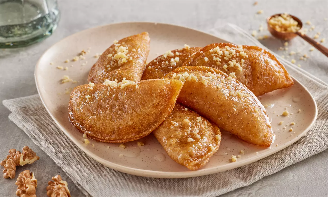 Zlatnosmeđi polumjesečasti tas kadayifi s hrskavom površinom, posuti mljevenim orasima, položeni na tanjur svijetle boje