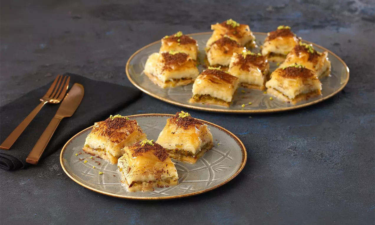 Kocke baklave s hrskavim zlatnosmeđim slojevima tijesta, zelenkastim tragovima pistacija, poslužene na tanjiru s tamnom podlogom