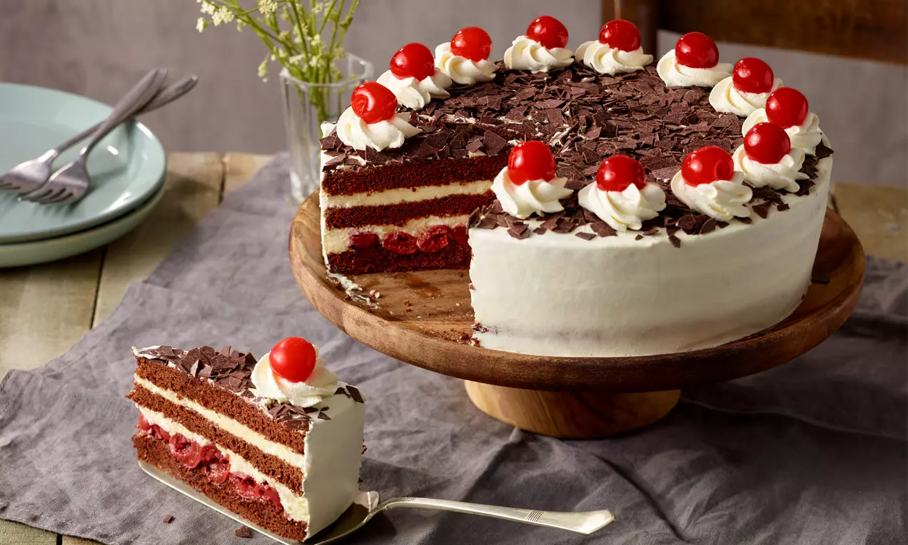 Torta s tamnim čokoladnim korama, slojevima crvenog voća i bijelog šlaga, ukrašena šlag rozetama, trešnjama i čokoladnim listićima