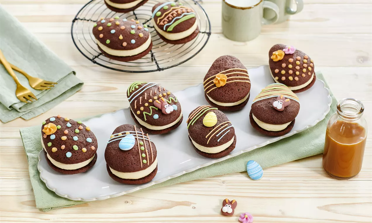 Čokoladni kolačići ovalnog oblika s bijelim krem punjenjem, ukrašeni šarenim glazurama i šećernim dekoracijama na pladnju od bijelog porculana