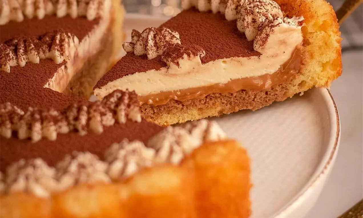 Torta s biskvitnom podlogom, slojem svijetle i tamnije kreme, ukrašena šlagom i kakaom, poslužena na bijelom tanjiru.