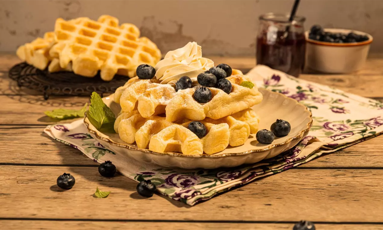 Četvrtasti svijetlozlatni vafli s neravnom teksturom, posuti šećerom, složeni na plavom tanjiru, ukrašeni šlagom i borovnicama
