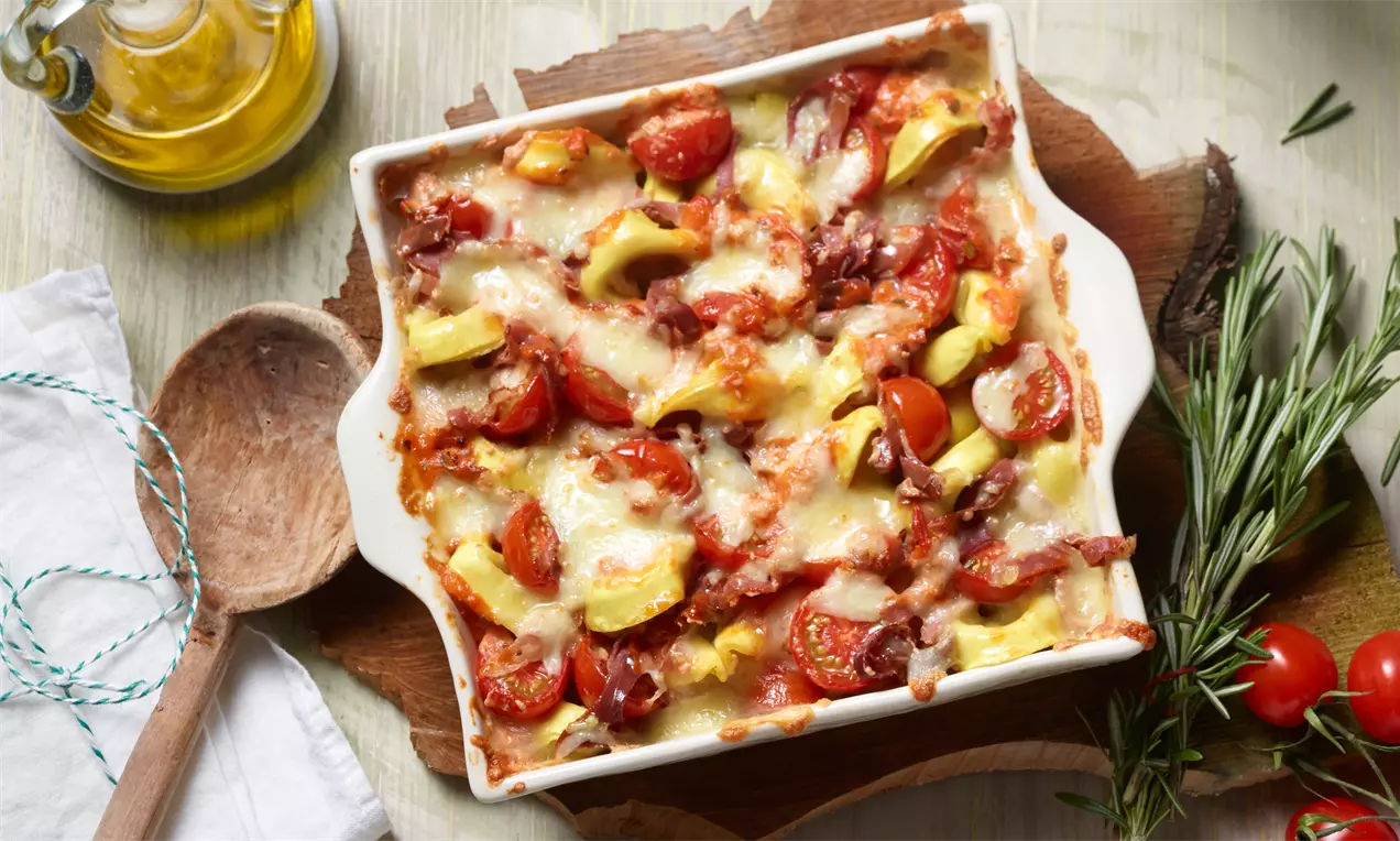 Pečeni tortelloni u bijeloj porcelanskoj posudi, prekriveni rastopljenim sirom, parmezanskom šunkom, kriškama tikvica i čeri rajčicama