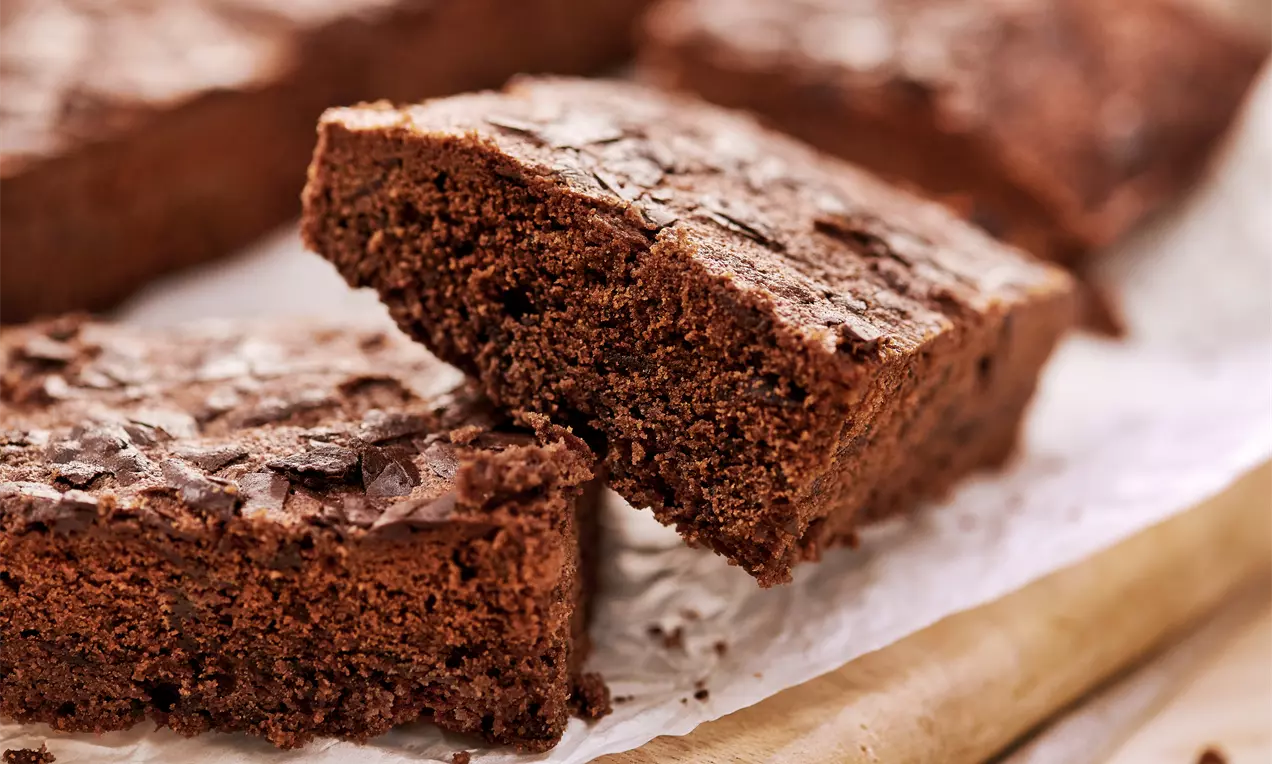 Čokoladni brownie kvadratnog oblika s hrskavom, ispucalom koricom na površini, naslagan na papiru za pečenje u rustikalnom okruženju