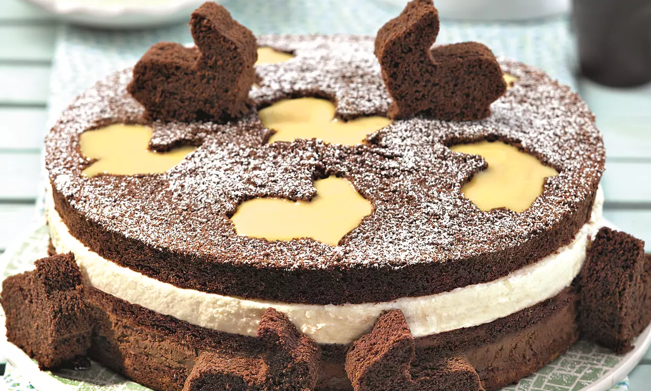 Čokoladna torta tamne smeđe boje s bijelim slojem kreme, rupičastim vrhom posutim šećerom u prahu i čokoladnim dekoracijama u obliku zečića