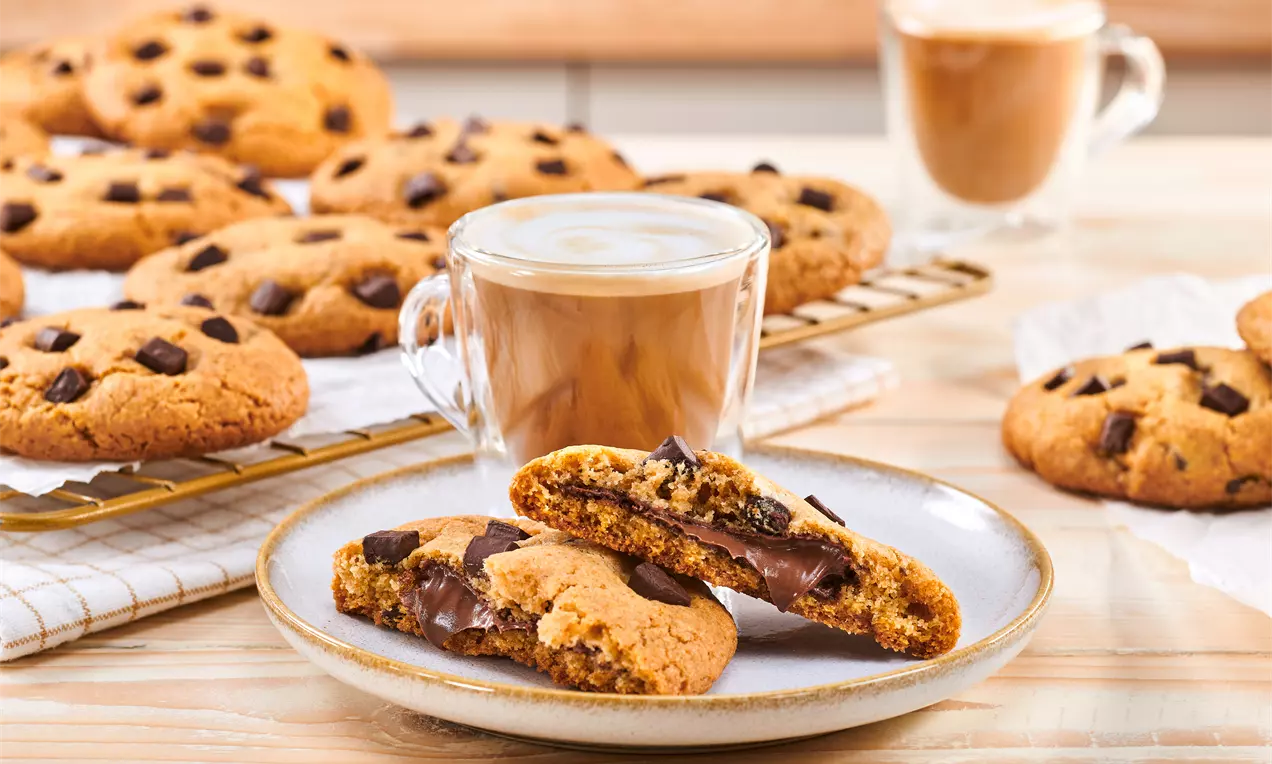 Zlatnosmeđi keksi s tamnim čokoladnim komadićima, jedan prerezan otkrivajući kremasto punjenje, na tanjuru uz staklenu šalicu kave