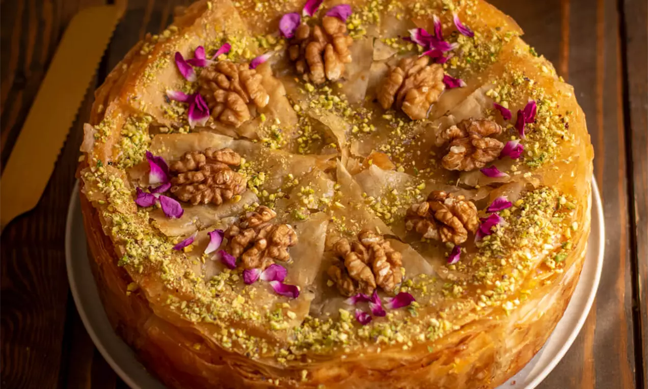 Okrugla torta od tankih zlatnosmeđih kora, posuta mljevenim pistaćima, ukrašena orasima i ljubičastim cvjetovima na bijelom tanjiru