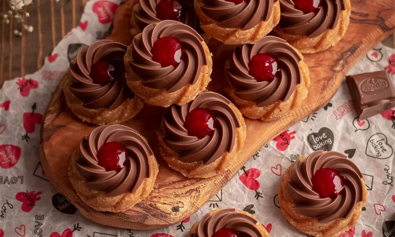 Zlatnosmeđi prhki kolačići kružnog oblika s kremom od čokolade i crvenom trešnjom u sredini, posluženi na drvenoj dasci