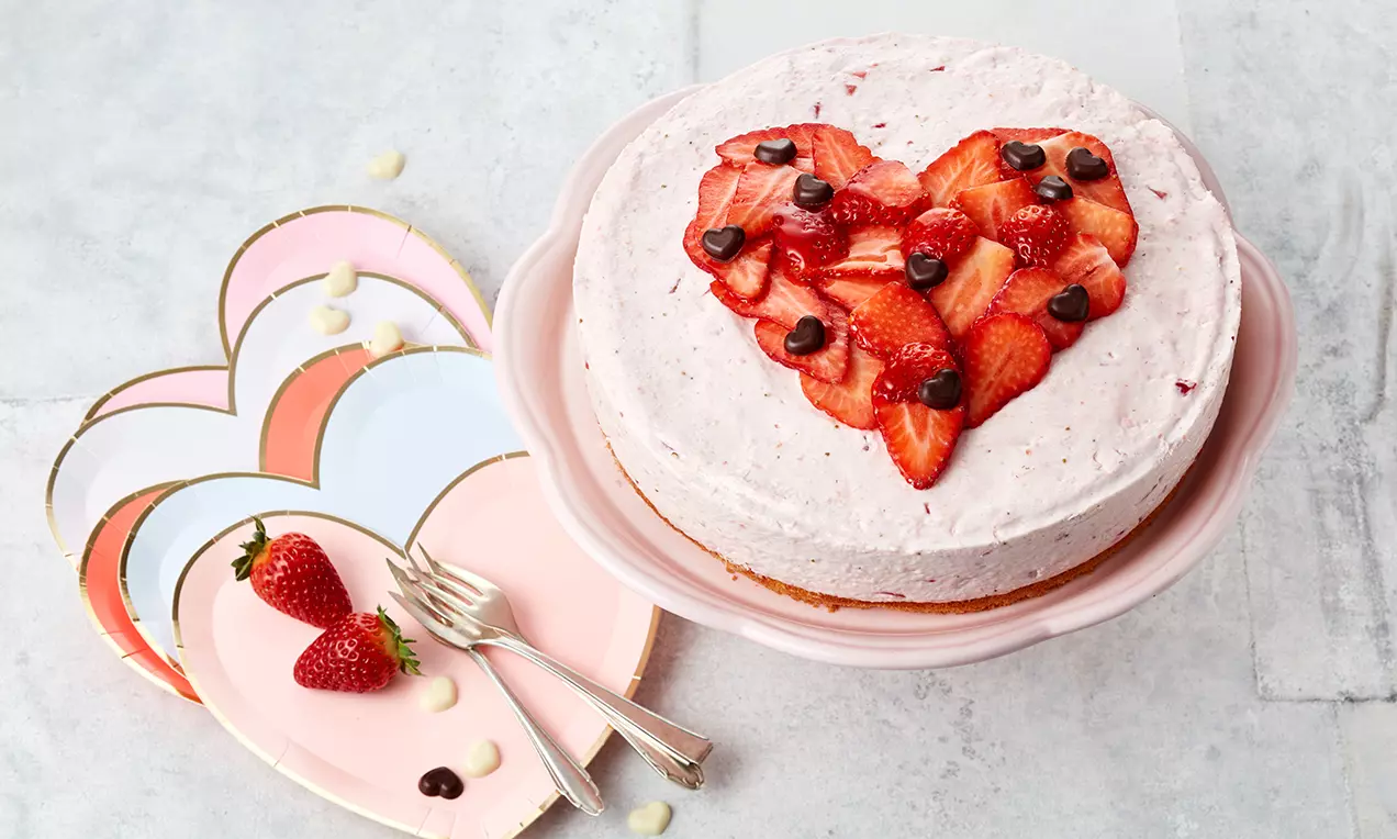 Torta svijetloružičaste kreme na biskvitnoj podlozi ukrašena jagodama u obliku srca i čokoladnim kapljicama na ružičastom tanjuru