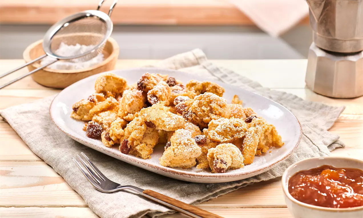 Na ovalnom tanjuru svijetlih tonova posute šećerom u prahu nalaze se zlatnosmeđi komadići Kaiserschmarrna uz zdjelicu džema.